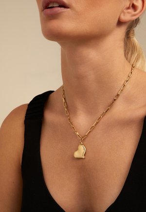 Femme portant une chaîne en or avec un pendentif en forme de cœur sur un haut sans manches noir, sur un fond beige.