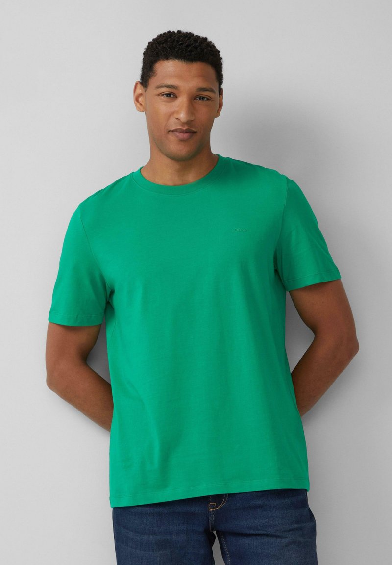 Zielona bawełniana koszulka z okrągłym dekoltem i krótkimi rękawami, z delikatnym logo na klatce piersiowej. Noszona z niebieskimi dżinsami.