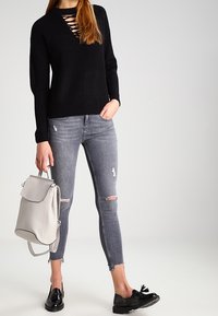 Jersey negro con escote cut-out, jeans skinny desgastados grises, zapatos de charol negros y una mochila de cuero gris claro.
