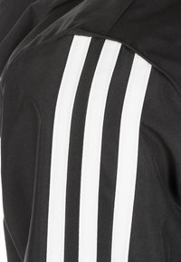 Czarna tkanina z trzema pionowymi białymi paskami, prawdopodobnie część ramienia sportowej kurtki lub bluzy.