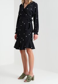 Robe portefeuille noire à pois blancs, manches longues, ourlet à volants et ceinture à nouer ; associée à des sandales à talons blocs nouées vert olive.