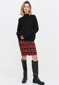 Czarny sweter z dzianiny, spódnica w czerwoną i czarną kratę oraz czarne kozaki do kolan. Stylizacja charakteryzuje się luźnym krojem i gładkimi fakturami.