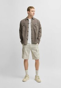 Lichtbruine linnen shirt met twee borstzakken, witte t-shirt eronder, beige cargoshorts en beige sneakers met dikke zolen.