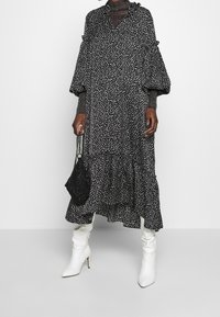 Robe noire à pois blancs, avec un ourlet en niveaux et des manches ballons. Associée à des bottes blanches à talons et un sac noir texturé.