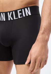 Κοντινό πλάνο στο κάτω μέρος του κορμού ενός άνδρα που φοράει μαύρα μπόξερ Calvin Klein με λογότυπο στη μέση και χαλαρό χέρι στο πλάι.