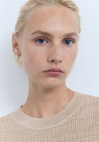 Beige strikket sweater med et struktureret mønster og en rund halsudskæring. Modellen har lyst blont hår og minimal smykker, med en enkel baggrund.
