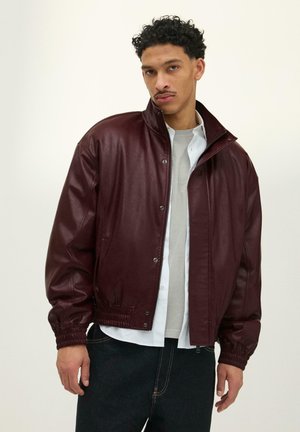 Mies, jolla on lyhyet kiharaiset hiukset, pukeutuneena tummanpunaiseen nahkatakkiin valkoisten ja harmaiden paitojen päällä, seisoo yksinkertaisen vaalean taustan edessä.