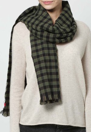 Femme portant une écharpe à carreaux verts et noirs enroulée autour du cou, par-dessus un pull beige et un pantalon foncé.