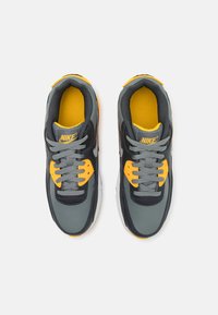 Nike ketsid on halli välisküljega, millel on mustad aksendid, kollased rõhuasetused keeles ja silmade juures ning hallid paelad. Materjal on tekstuuritud ja nähtav pehmendus.