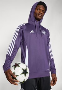 Violetti kevyt huppari valkoisilla yksityiskohdilla, jossa on vetoketjullinen etuosa, Adidas-logo ja Manchester Unitedin vaakuna. Musta jalkapallo, jossa on tähtikuvio.