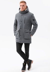 Grå vinterjacka med huva, som har två framfickor och en ficka på bröstet med dragkedja. Buren över svarta jeans och svarta kängor.