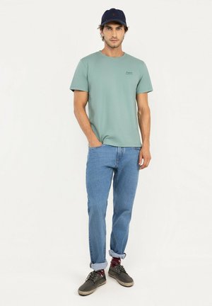 Hombre con camiseta verde azulado, vaqueros azules remangados, zapatillas oscuras, calcetines burdeos y una gorra azul marino, de pie con una mano en el bolsillo.