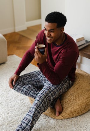 Chemise rouge à manches longues avec accents bleu marine, pantalon de pyjama à carreaux bleus et gris, assis sur un pouf tressé, tenant une tasse avec une cuillère.