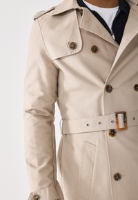 Trench coat beige hecho de tela suave, con grandes botones marrones, cintura ceñida con cinturón y un frente de doble botonadura con un cuello con muesca.
