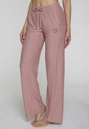 Frau trägt pinke Lounge-Hose mit Kordelzug und "Cozy Athleisure"-Patch, barfuß, steht vor einem schlichten, hellen Hintergrund.