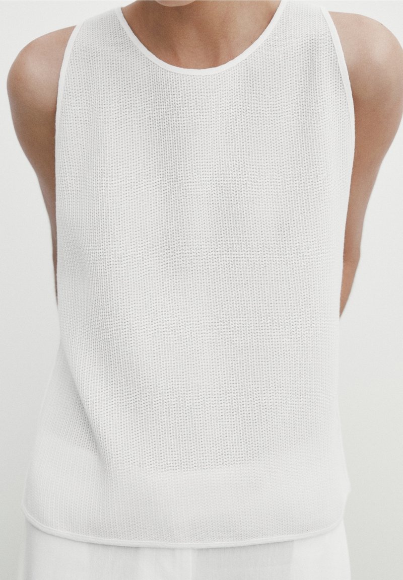 Haut blanc sans manches en tissu maille texturé, avec un col rond et une coupe décontractée, légèrement plus longue à l'arrière.