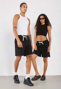 Juoda sportinė šortai su balta juostele, balta įmantri tank top marškiniai ir juodas įmantris cropped marškinėlis, derinami su juodais sportiniais bateliais ir šlepetėmis.