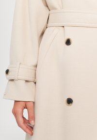 Beige Wollemantel mit tailliertem Gürtel, großen Knöpfen und umgeschlagenen Ärmeln. Der Stoff ist weich und strukturiert und hat eine glatte Oberfläche.