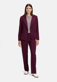 Kobieta w ciemnofioletowym garniturze, w paskowaną bluzkę i białych sneakersach, stojąca na jednolitym jasnym tle.