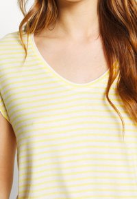 Žluté a bílé pruhované tričko s jemnou texturou, se širokým výstřihem a krátkými rukávy. Tkanina vypadá lehce a ležérně.