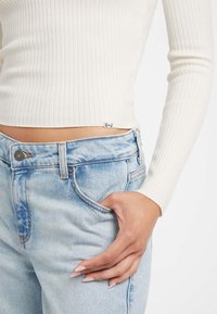 Mano nella tasca dei jeans di denim azzurro chiaro, indossando un crop top a maniche lunghe bianco a costine, che mostra il ventre e le unghie curate.