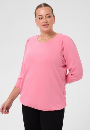 Kvinde iført en lyserød bluse med lange ærmer og sorte bukser, med håret trukket tilbage og minimal smykker, mod en ensfarvet baggrund.