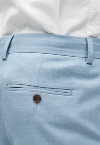 Ανοιχτό μπλε παντελόνι με πίσω τσέπη με κουμπί και θηλιές για ζώνη, φορεμένο με λευκό πουκάμισο μέσα από το παντελόνι.