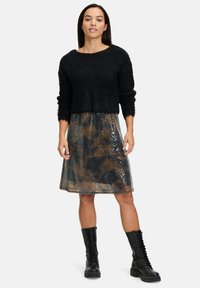 Schwarzer, verkürzter Pullover mit strukturiertem Strick, kombiniert mit einem semi-transparenten, bedruckten Rock mit Pailletten. Das Modell trägt schwarze Schnürstiefel.