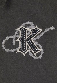 Black fabric patch features a stylized letter "K" adorned with rhinestones, surrounded by decorative silver stitching and accents.