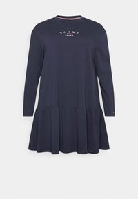 Granatowa sukienka z długimi rękawami o warstwowym dole, z okrągłym dekoltem i białym logo "TOMMY JEANS" na piersi. Gładka bawełniana tekstura.