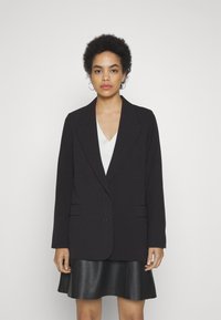 Blazer noir ajusté, avec des revers crantés et deux poches avant, associé à un haut blanc à col en V et une jupe en cuir noir.