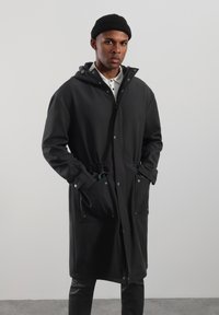 Czarny długi płaszcz z kapturem, zapinany na zatrzaski, z dwoma przednimi kieszeniami i regulowanym sznurkiem w talii. Gładka tekstura.