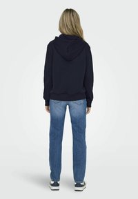 Felpa navy con cappuccio, polsini e vita a coste. Abbinata a jeans azzurri chiari e sneaker. La vista posteriore mostra una vestibilità comoda e uno stile informale.