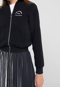 Schwarze Bomberjacke aus glattem Stoff, mit Reißverschluss, gerippten Bündchen und gesticktem Logo. Kombiniert mit einem plissierten schwarzen Rock.