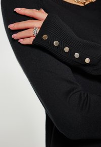Pulover negru tricotat, cu manșete ribbed decorate cu cinci nasturi metalici. Prim-plan al detaliilor mâinii și manșetei.