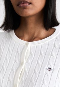 Witte kabelgebreide cardigan met ribbelranden, een ronde hals en een klein geborduurd logo aan de linkerkant. Knopsluiting zichtbaar.