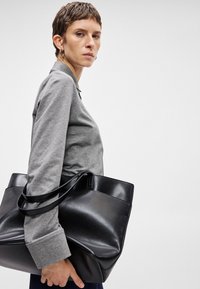 Μαύρη δερμάτινη τσάντα tote με διπλές λαβές, δομημένο σχήμα, υφή επιφάνειας και λεπτομέρεια μικρής τσέπης. Φοριέται από ένα άτομο που φοράει γκρι τοπ.