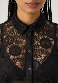 Μαύρη μπλούζα από δαντέλα με πολύπλοκα λουλουδάτα σχέδια, γιακά και κλείσιμο με κουμπιά στο μπροστινό μέρος. Υφή από δαντέλα με διαφανή εφέ.