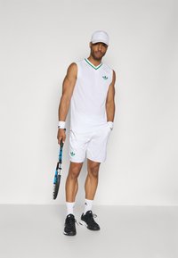 Weißes ärmelloses Sporttop mit grünen Akzenten und Logo, kombiniert mit weißen Shorts. Schwarze Sportschuhe, hält einen Tennisschläger.