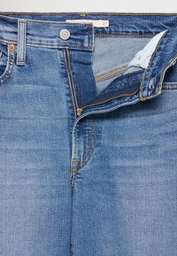 Jean en denim bleu avec une fermeture éclair ouverte, un bouton argenté et une étiquette de taille 27 à la ceinture.