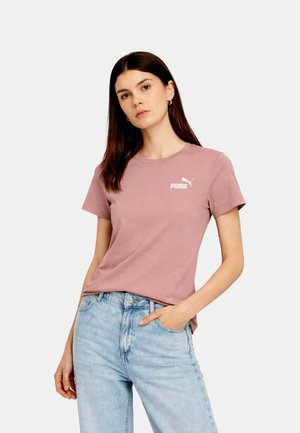Donna che indossa una t-shirt mauve Puma e jeans azzurri, in piedi con una mano in tasca davanti a uno sfondo chiaro e semplice.