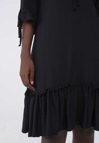 Svart klänning med en volangkjol, lättviktsmaterial och långa ärmar med knytakenter. Slät yta utan synliga mönster.