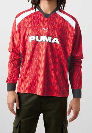 Mężczyzna w czerwonej koszuli Puma z długim rękawem, z białymi akcentami na ramionach i czarnym kołnierzem, w ciemnych spodniach cargo.