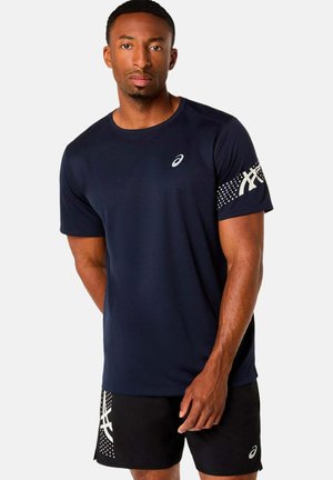 Mann trägt marineblaue ASICS-T-Shirt und schwarze Shorts mit weißem Logo und Punktmustern, steht vor einfachem Hintergrund.