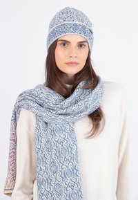 Donna che indossa un cappello di lana a maglia blu e bianco con un motivo abbinato e una sciarpa coordinata sopra un maglione color crema, rivolta in avanti con un'espressione neutra.