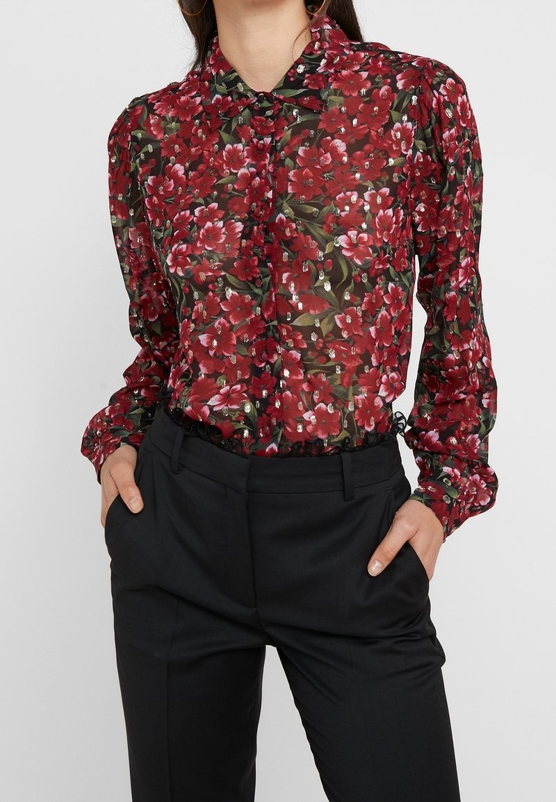 Blusa trasparente rossa a fiori con maniche lunghe, chiusura con bottoni davanti e polsini arricciati. Abbinata a pantaloni neri su misura per un contrasto.