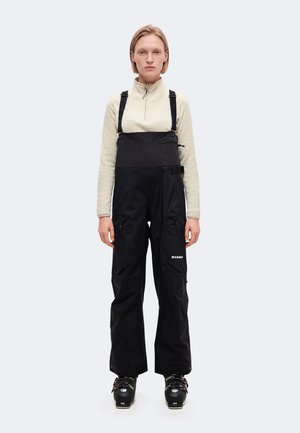 Un mannequin se tient devant un fond uni, vêtu d’un pantalon de ski noir à bretelles, d’un haut en polaire crème et de chaussures de ski.