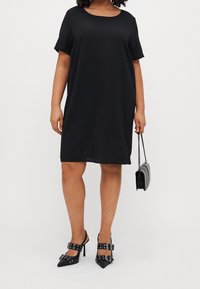 Vestido negro de manga corta con escote redondo, corte holgado y textura suave. Combinado con zapatos de tacón negro con hebillas decorativas.