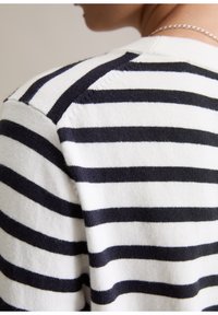 Gros plan d’une personne portant un pull blanc à rayures noires horizontales et un collier en chaîne argentée, montrant le haut du dos et la zone des épaules.