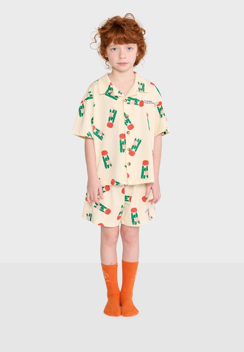 Enfant aux cheveux roux bouclés porte un t-shirt crème à manches courtes et un short avec des motifs abstraits verts et rouges, assortis à des chaussettes orange.